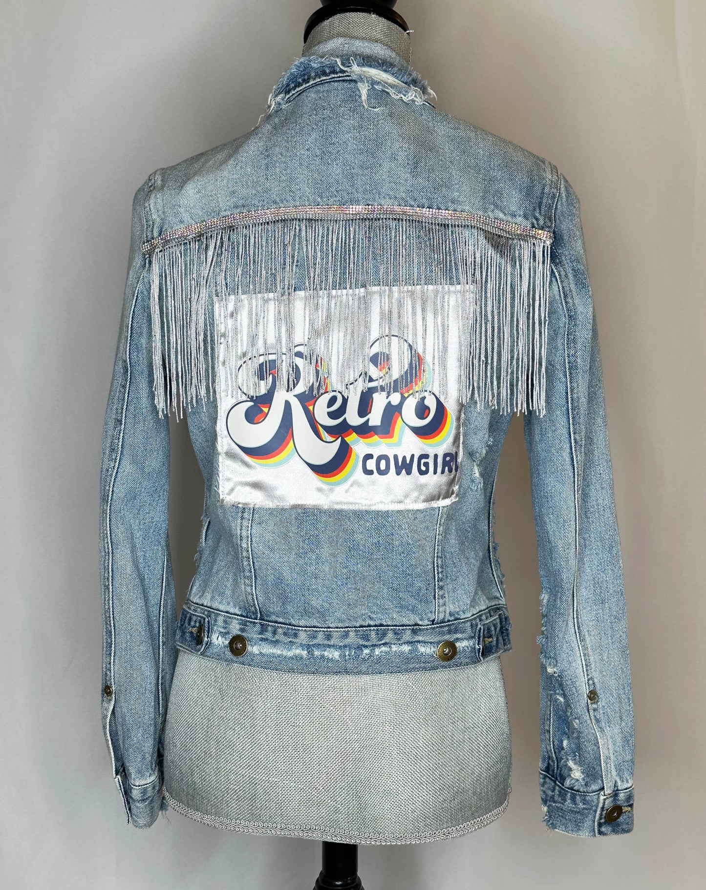 "Retro Cowgirl" Women's Denim jacket Back Side featuring rhinestone and fringe detailing with satin patch and double button detailing at the bottom.